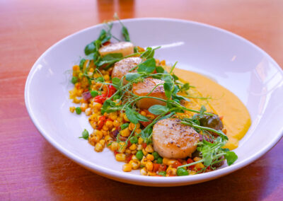 Seared Scallops Four seared scallops placed over cherry tomaroes, red peppers, corn, bacon, peas, corn puree, and pea tendrils. A pretty delicate dish with lots of color and vibrancy.